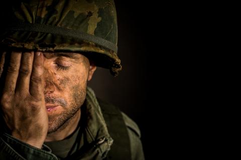 A dirt covered soldier wearing a helmet and covering his face with his hand.