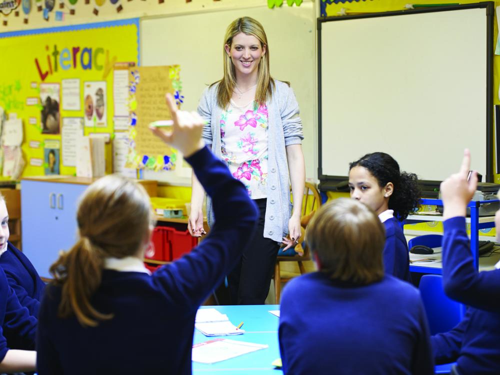 Female teacher with long blonde hair, blue sweater and pink blouse stands in front of a classroom of students with their arms raised.