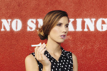 Photo of a woman smoking a cigarette while standing in front of a "no smoking" sign.
