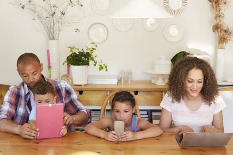 Photo of a family sitting at the table, all with electronic devices in front of them.