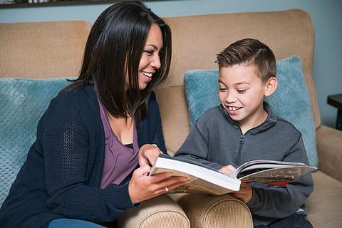 Mom and son looking at a book together smiling while sitting on a tan couch with blue pillows.