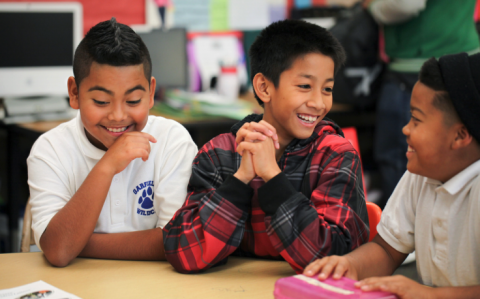 Photo of three children sitting at a classroom table and laughing.