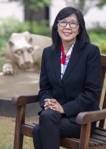 Karen Kim sitting down in chair outside Nittany Lion Shrine in black suit, glasses, long black hair and red, white, and blue scarf.