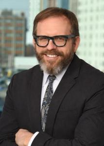 Headshot of Craig Newschaffer with brown hair, beard, glasses, white shirt, gray swirled tie, and black jacket.
