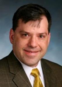 Headshot of Gregory Shearer with short black hair, white shirt, gold tie, and brown jacket.