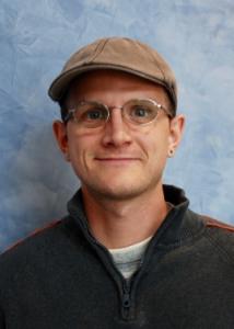 Headshot of Kyle Husmann with brown hat, glasses, white shirt, and gray jacket.
