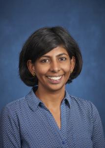 Headshot of Maithreyi Gopalan with mid-length, dark hair wearing a blue shirt in front of a blue background. 