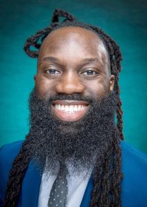 Headshot of Wilson Okello, a black man with a long beard, blue blazer, and teal and white patterned tie.