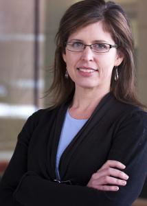 Headshot of Rachel Smith with long brown hair parted in the middle, black wire rimmed glasses, black jacket, and light blue shirt.