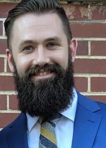 Headshot of Zachary Smith with short brown hair and long brown beard with blue suit, white shirt, and striped tie.